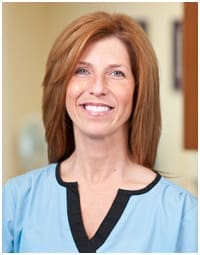 Gasior Dental Ann Arbor A woman with straight, shoulder-length auburn hair smiles at the camera. She is wearing a light blue top with a black-trimmed neckline, standing indoors. This Ann Arbor dentist is captured in a welcoming, professional atmosphere.