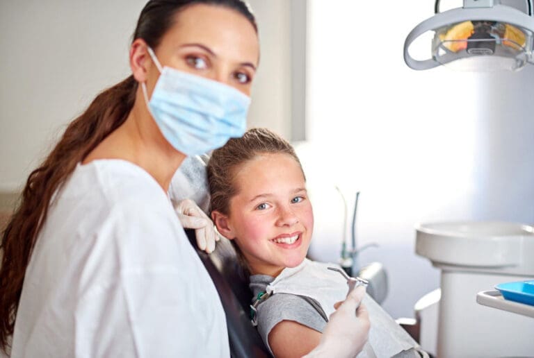 Gasior Dental Ann Arbor A dentist wearing a mask and gloves sits beside a smiling young girl in a dental chair. The girl holds a dental tool, and dental equipment is visible in the background.